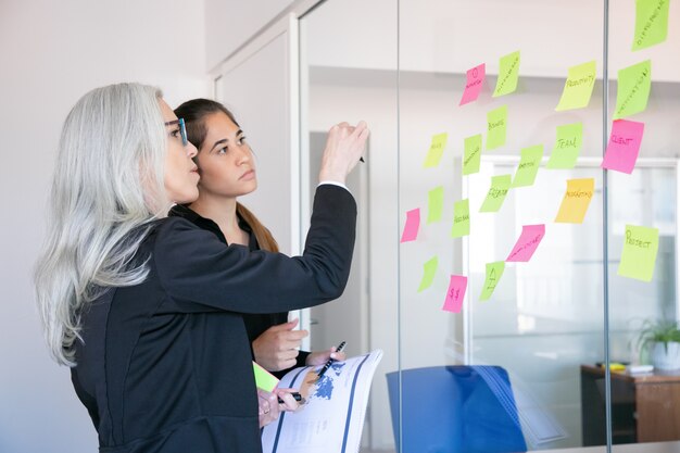 Empresarias concentradas mirando pegatinas en la pared de vidrio. Trabajadora de pelo gris concentrada tomando notas para la estrategia o plan del proyecto