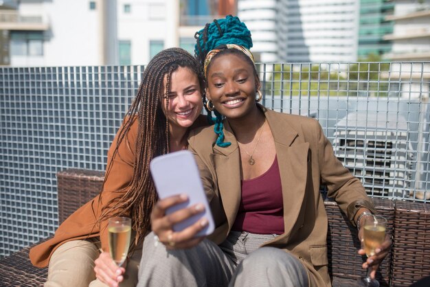 Empresarias felices haciendo selfie en fiesta en la azotea de la terraza. Mujeres en ropa formal sentado en un sofá de ratán, mirando el teléfono móvil, sonriendo. Teambuilding, fiesta, concepto de redes sociales