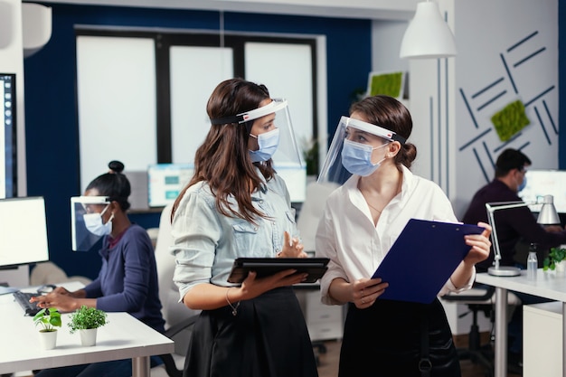 Empresarias con máscaras de protección contra el coronavirus de pie en el lugar de trabajo hablando de los datos financieros de la empresa con tableta digital. Equipo de negocios multiétnico trabajando respetando la distancia social