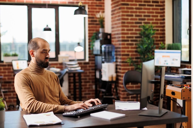 Empresario adulto joven que desarrolla ideas financieras y gráficos de gestión. Feliz y sonriente oficinista profesional mirando gráficos financieros mientras se sienta en el escritorio usando una computadora de trabajo.