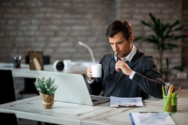 Empresario adulto medio usando computadora mientras trabaja en informes y tomando café en el trabajo