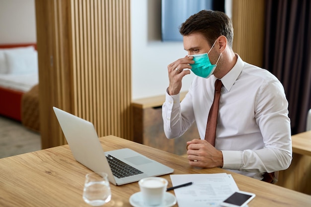 Empresario ajustando mascarilla protectora mientras trabaja en una computadora en la habitación del hotel