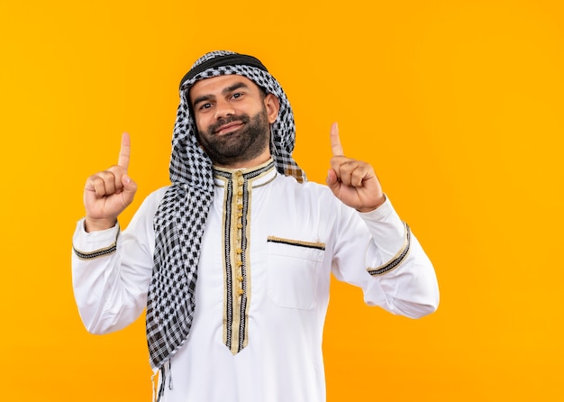 Foto gratuita empresario árabe en ropa tradicional apuntando con el dedo índice hacia arriba sonriendo confiado de pie sobre la pared naranja