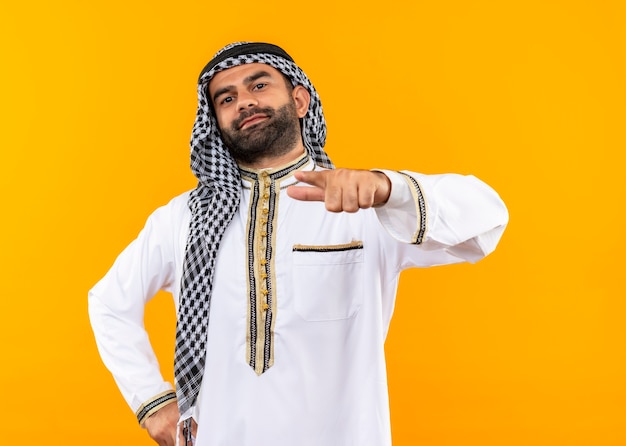 Foto gratuita empresario árabe en ropa tradicional apuntando con el dedo sonriendo confiado de pie sobre la pared naranja