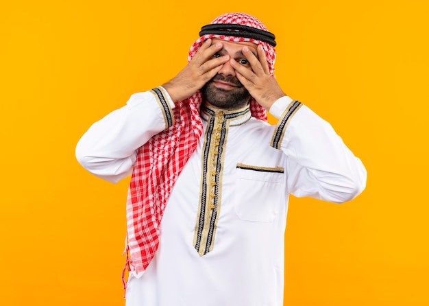 Empresario árabe en ropa tradicional cerrando los ojos con las palmas mirando a través de los dedos de pie sobre la pared naranja