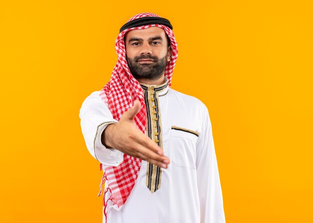 Empresario árabe en ropa tradicional mirando confiado saludo ofreciendo mano de pie sobre la pared naranja
