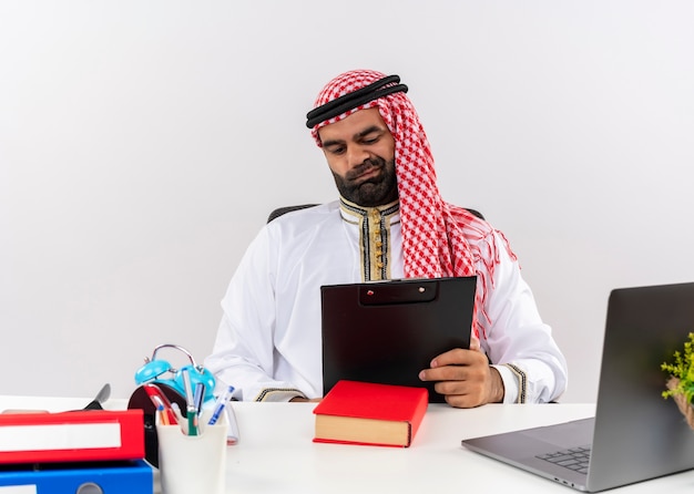 Empresario árabe en ropa tradicional sentado en la mesa con portapapeles mirando documentos disgustado trabajando en la oficina