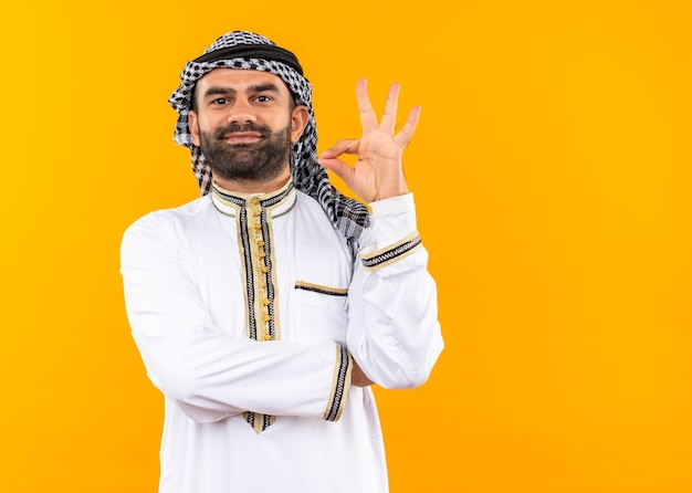 Empresario árabe en ropa tradicional sonriendo confiado haciendo bien firmar de pie sobre la pared naranja