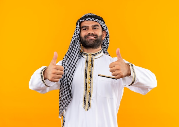 Foto gratuita empresario árabe en ropa tradicional sonriendo confiado mostrando thumbs up parado sobre pared naranja