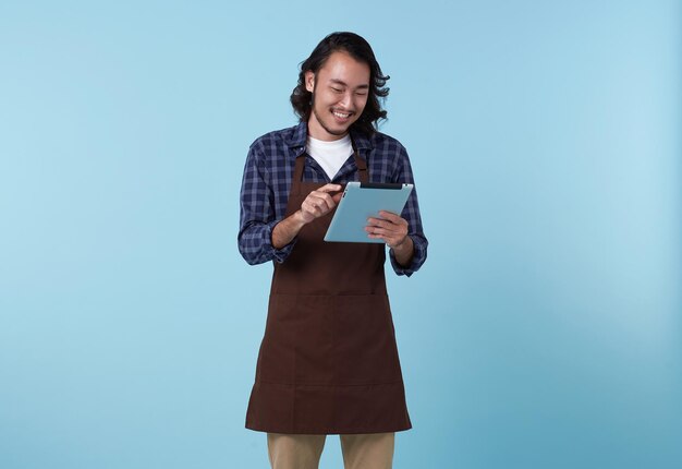 empresario asiático hombre en delantal mano sujetando tableta para listar comida de pedido aislado sobre fondo azul