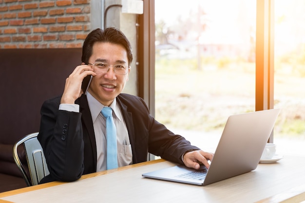 Empresario asiático usando laptop en cafetería hablando por teléfono