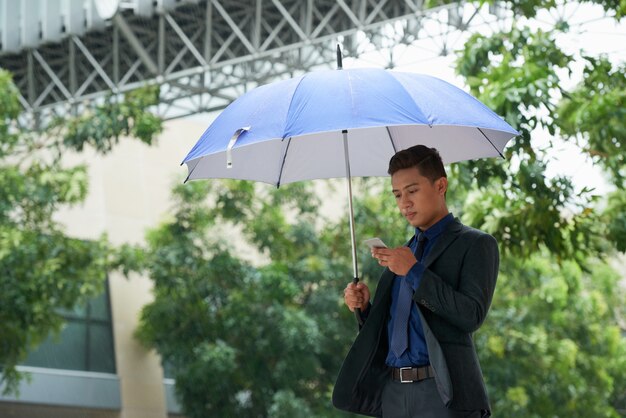 Empresario chino de pie con paraguas bajo la lluvia y con smartphone