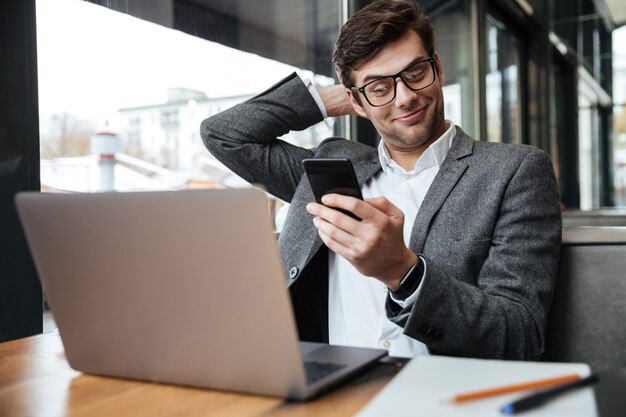 Empresario complacido en anteojos sentado junto a la mesa en la cafetería con computadora portátil mientras usa el teléfono inteligente