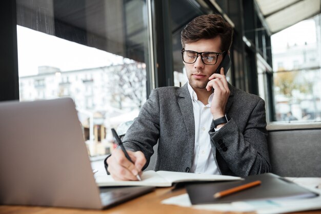 Empresario concentrado en anteojos sentado junto a la mesa en la cafetería con computadora portátil mientras habla por teléfono inteligente y escribe algo