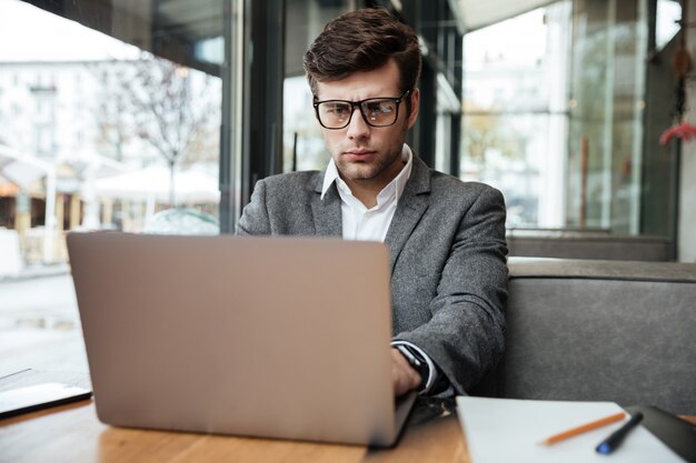 Empresario concentrado en anteojos sentado junto a la mesa en la cafetería y usando la computadora portátil