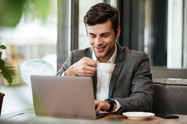 Empresario contento sentado junto a la mesa en la cafetería mientras sostiene una taza de café y usa la computadora portátil