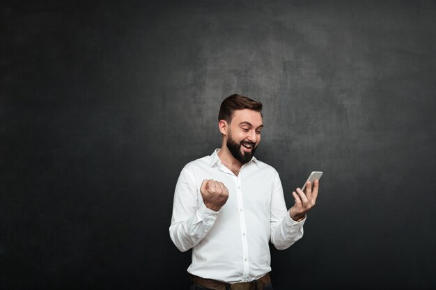 Empresario encantado en camisa blanca actuando como ganador apretando el puño mientras usa el teléfono celular sobre grafito