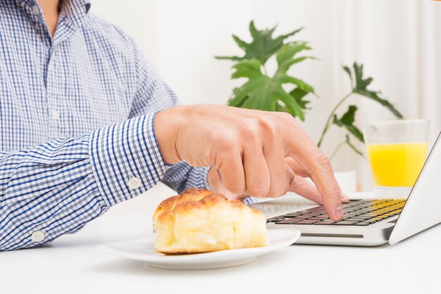 Empresario escribiendo en la computadora portátil con pan y vaso de jugo en la mesa