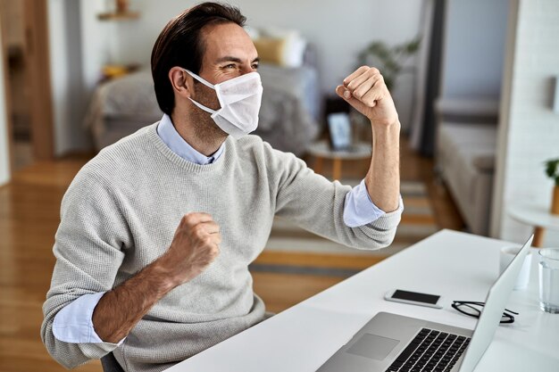 Empresario feliz con mascarilla protectora celebrando el éxito mientras trabaja en una computadora en casa