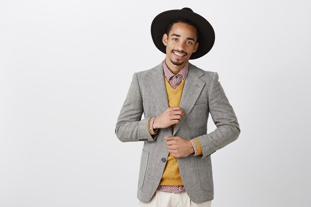 Empresario de fiesta, celebrando un trato exitoso. Confiado y encantador empresario afroamericano con elegante ropa formal y sombrero, revisando el atuendo y sonriendo ampliamente, siendo descarado sobre la pared gris