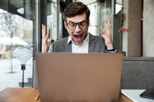 Empresario gritando sorprendido en anteojos sentado junto a la mesa en la cafetería y regocijarse mientras mira la computadora portátil