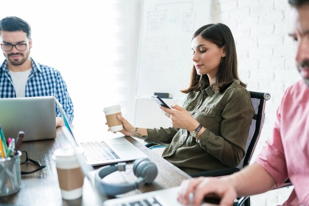 Empresario hispano socializando en un teléfono inteligente mientras toma un café en el escritorio de la oficina