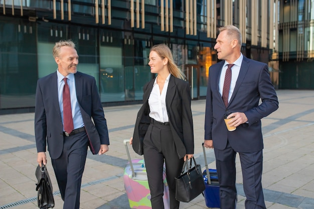 Empresario líder en colegas extranjeros. Empresarios caminando en el edificio de oficinas, maletas con ruedas, hablando. Concepto de viaje de negocios