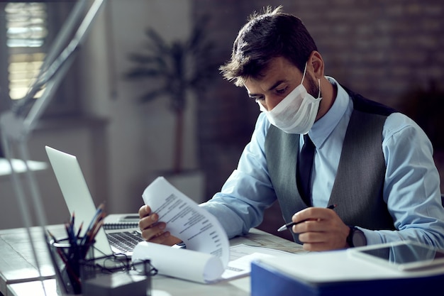 Empresario masculino analizando informes comerciales mientras usa mascarilla y trabaja en la oficina durante la epidemia de virus