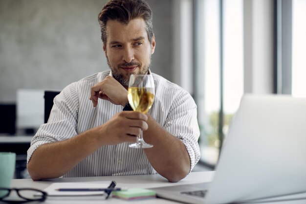 Empresario de mediana edad brindando con una copa de vino mientras realiza una videollamada a través de una laptop en la oficina