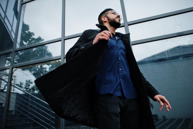 Empresario de oriente medio usa abrigo negro y anteojos de camisa azul contra el edificio de oficinas fumando cigarrillos