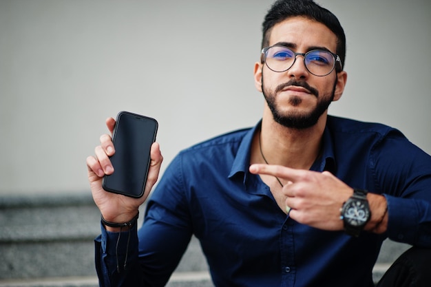 Foto gratuita empresario de oriente medio usa anteojos de camisa azul contra el edificio de oficinas sentado en las escaleras y muestra el dedo al teléfono