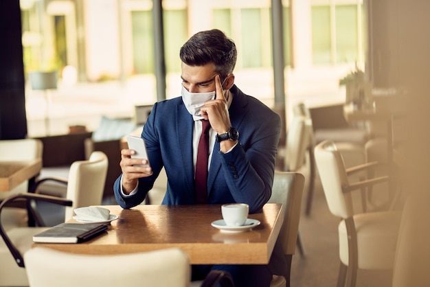 Empresario pensativo con mascarilla leyendo un mensaje de texto por teléfono en un café