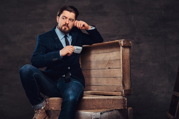 Empresario pensativo de mediana edad con cabello elegante y barba vestido con jeans, chaqueta y corbata, sentado en cajas de madera y sosteniendo una taza de café en un estudio. Aislado en un fondo de textura oscura.