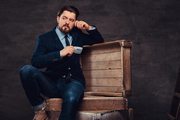 Empresario pensativo de mediana edad con cabello elegante y barba vestido con jeans, chaqueta y corbata, sentado en cajas de madera y sosteniendo una taza de café en un estudio. Aislado en un fondo de textura oscura.