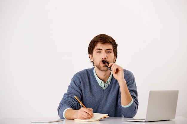 Empresario pensativo sentado en el escritorio de la oficina, escribiendo, tratando de pensar una idea, entrecerrando los ojos mientras reflexiona sobre el nuevo contenido