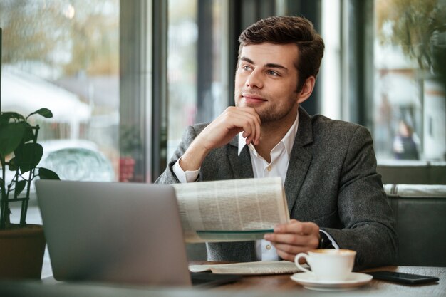 Empresario pensativo sonriente sentado junto a la mesa en la cafetería con computadora portátil y periódico mientras mira lejos