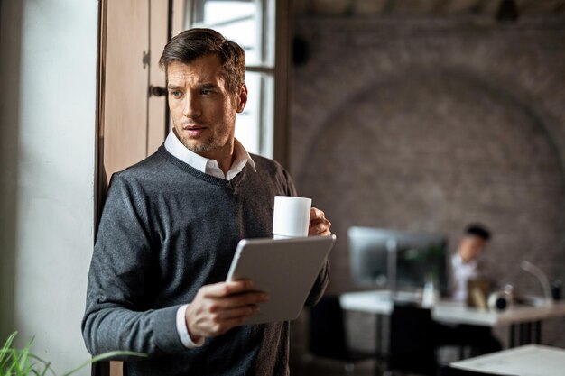 Empresario pensativo tomando café y usando el panel táctil mientras está parado en la oficina y mira hacia otro lado