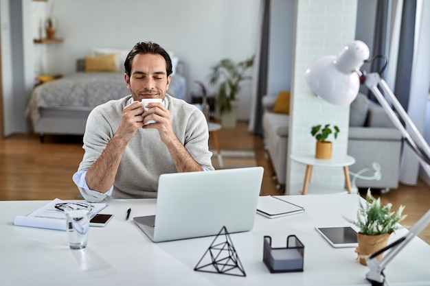 Empresario sonriente disfrutando de un descanso para tomar café con los ojos cerrados mientras trabaja en casa