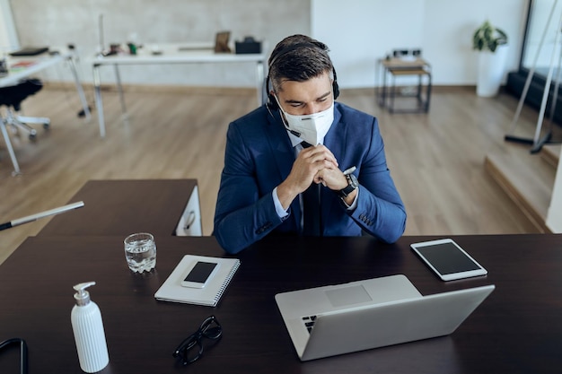 Empresario sonriente con máscara facial usando computadora mientras hace una videollamada en la oficina