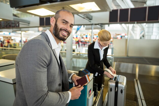 Empresario sonriente de pie con pasaporte mientras el asistente se pega la etiqueta al equipaje