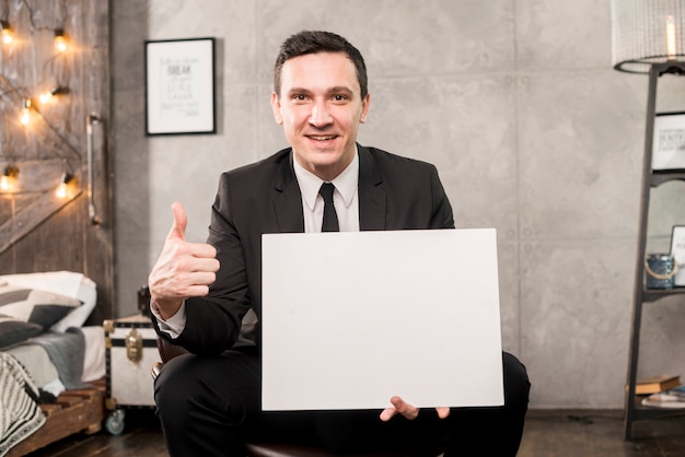 Empresario sonriente sosteniendo papel en blanco y gesticulando pulgar arriba