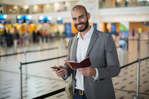 Empresario sonriente sosteniendo una tarjeta de embarque y comprobando su teléfono móvil