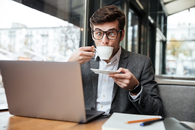Empresario sorprendido en anteojos sentado junto a la mesa en la cafetería con computadora portátil mientras bebe café y mira