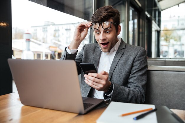 Empresario sorprendido en anteojos sentado junto a la mesa en la cafetería mientras sostiene el teléfono inteligente y mira la computadora portátil