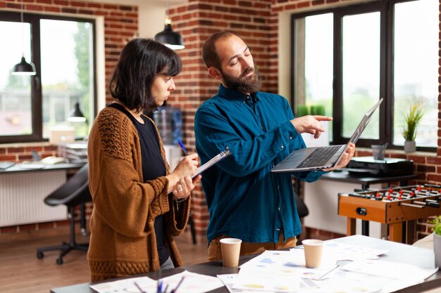 Empresario sosteniendo una computadora portátil que muestra estadísticas de marketing a una mujer gerente que trabaja en la estrategia de la empresa en la oficina de inicio. Colegas analizando el informe de gestión. Concepto de negocio