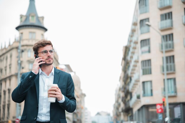 Empresario sosteniendo de pie en la calle sosteniendo una taza de café para llevar en la mano hablando por teléfono móvil