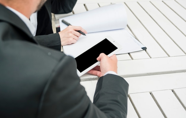 Empresario sosteniendo teléfono inteligente en la mano sentado con su colega sosteniendo la pluma sobre el documento