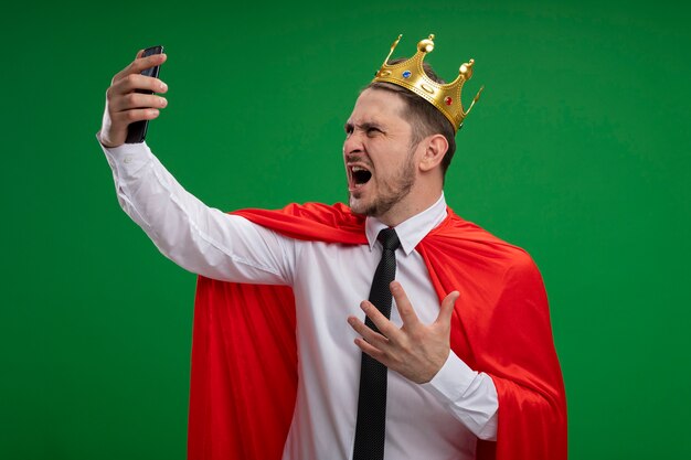 Empresario de superhéroe en capa roja con corona haciendo selfie con smartphone volviendo loco enojado de pie sobre fondo verde