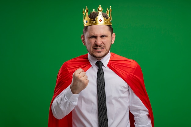 Empresario de superhéroe en capa roja con corona mirando a la cámara con cara enojada apretando el puño de pie sobre fondo verde