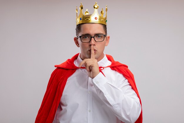 Empresario de superhéroe en capa roja y gafas con corona haciendo gesto de silencio con el dedo en los labios de pie sobre la pared blanca